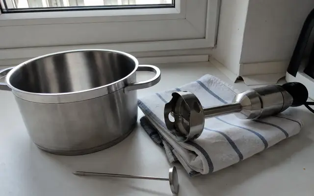 Pusty garnek ze stali, blender ręczny i termometr szpilkowy na białym blacie przy oknie